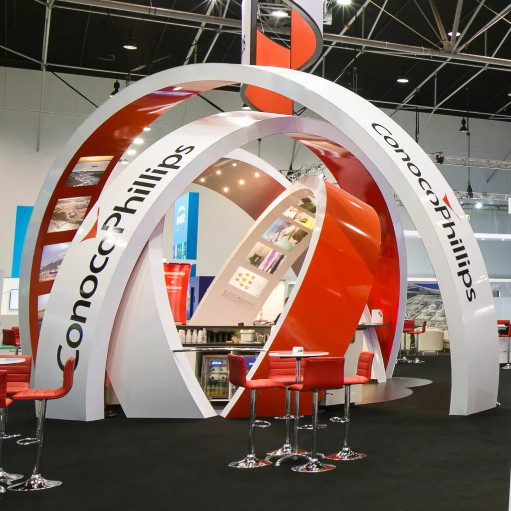 Modern exhibition booth with large, curved white and red arches featuring "ConocoPhillips" text. Red chairs and stools surround sleek tables on a black carpet.