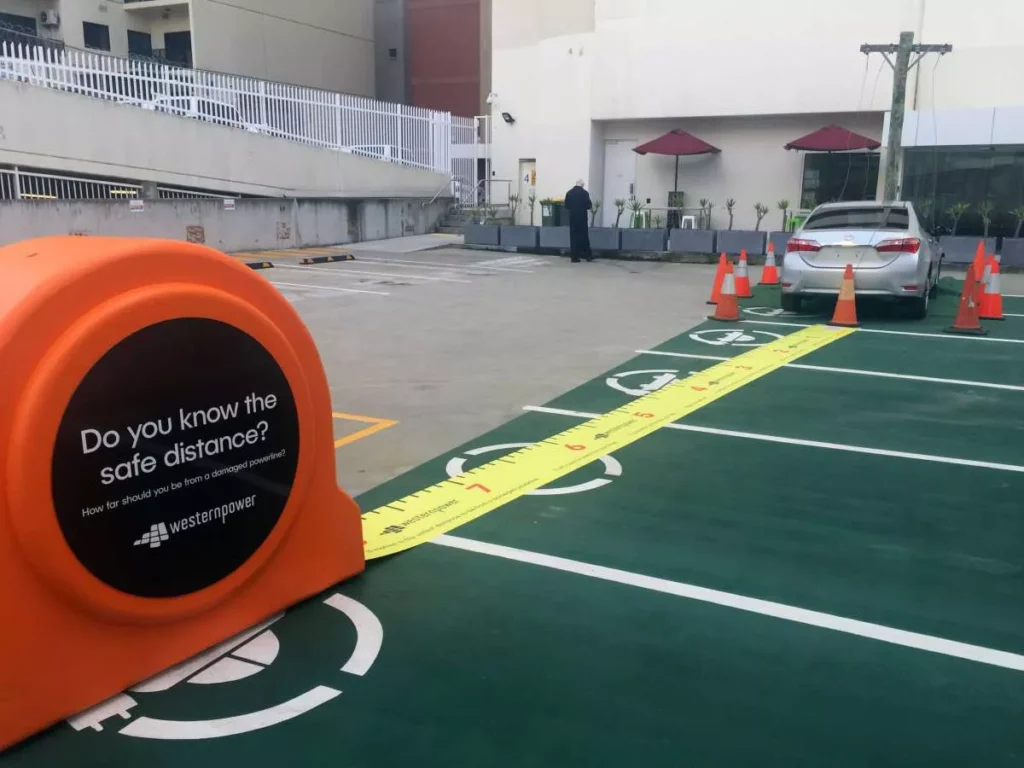 A large orange tape measure display on a parking lot shows safe distance guidelines. A silver car is parked nearby, with traffic cones outlining its space.
