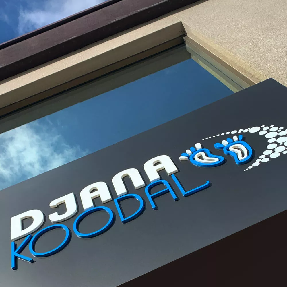 Sign with "DJANA KOODAL" in white and blue 3D letters on a dark background, featuring a pattern of stylized footprints