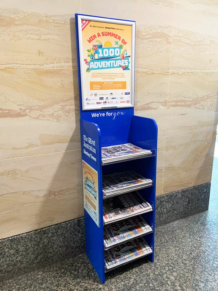 A blue informational display stand holds multiple rows of brochures titled "1,000 Adventures." It's positioned against a marble wall.