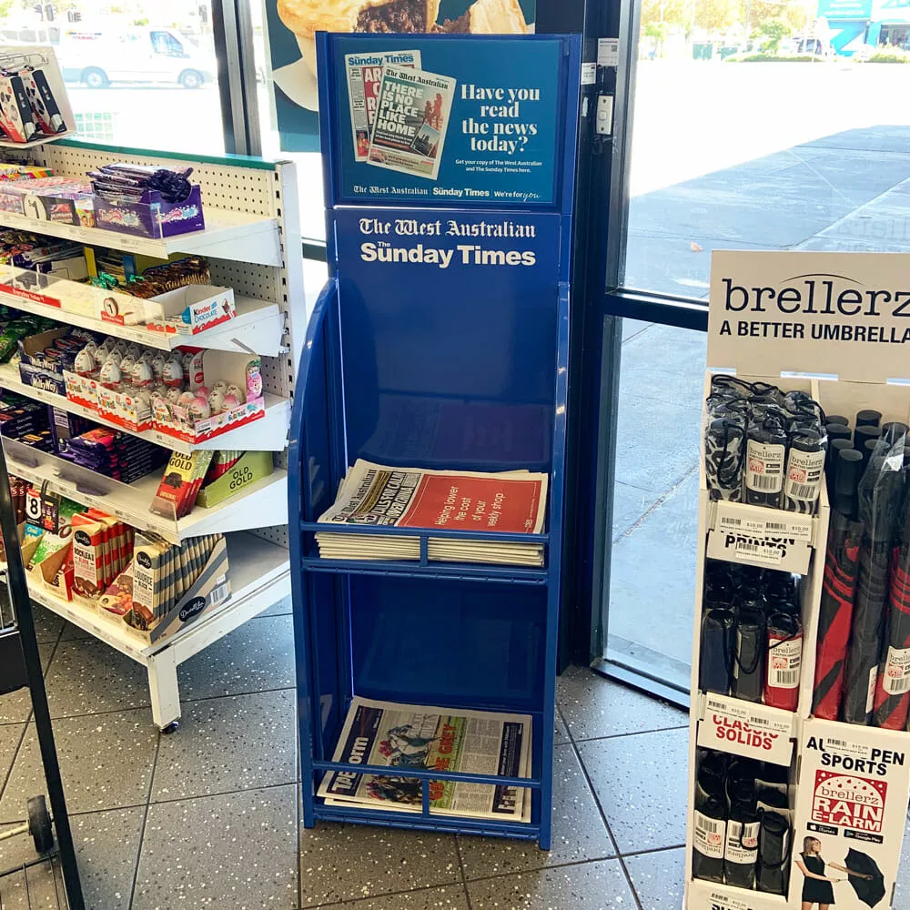 Blue newspaper stand for "The West Australian Sunday Times" is inside a store with shelves of snacks and umbrellas nearby.
