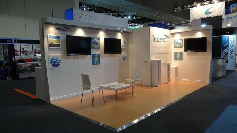 Exhibition booth with branding on walls, three flat screens, white chairs, and a small table on a wooden floor. Bright, modern, and inviting.