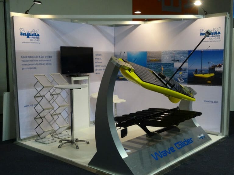 A trade show booth displays a Wave Glider model on a stand, with a screen and brochure stand nearby. The backdrop shows maritime images.
