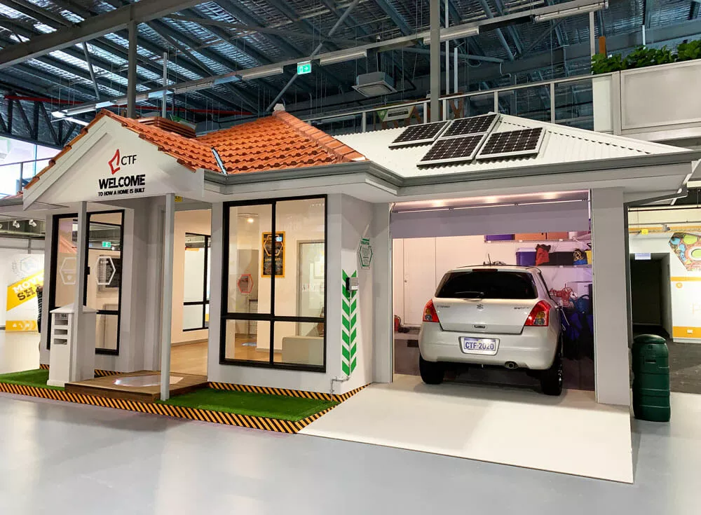 Miniature house model with solar panels on the roof and a car parked in a garage. Features a welcoming sign, large windows, and a realistic facade.