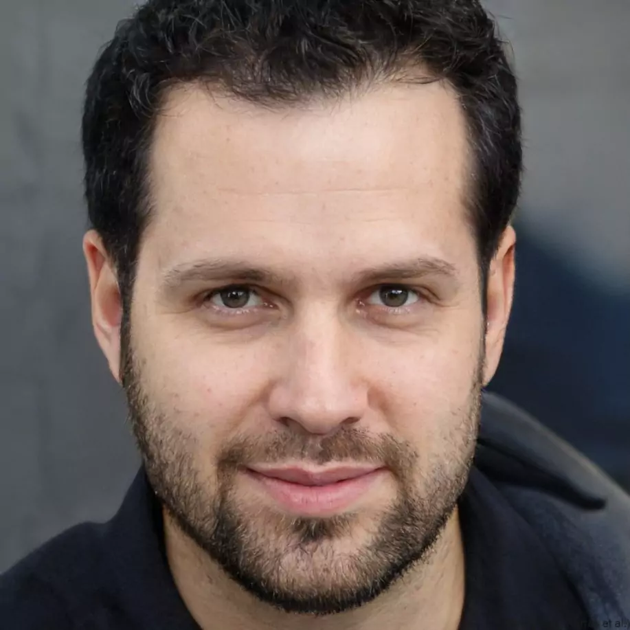 A man with dark hair and a beard smiles softly, conveying warmth and friendliness. He wears a black jacket against a blurred dark background
