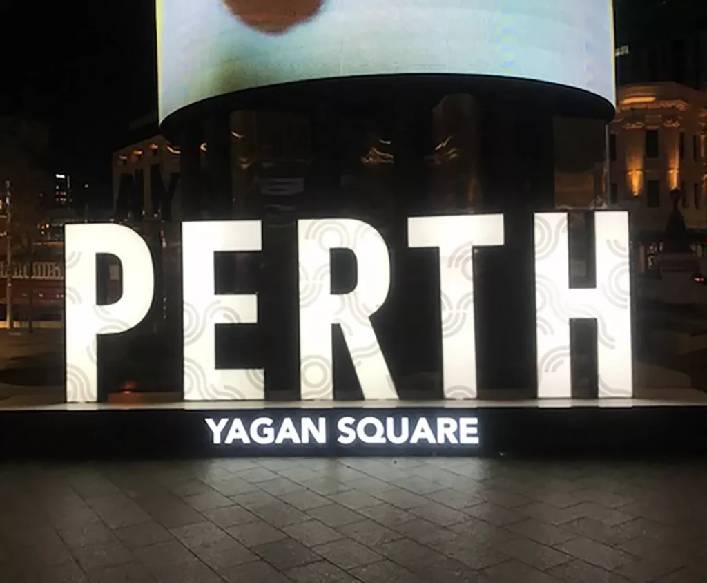 Illuminated "PERTH" letters with decorative patterns stand prominently in Yagan Square at night, with a vibrant urban atmosphere visible in the background.