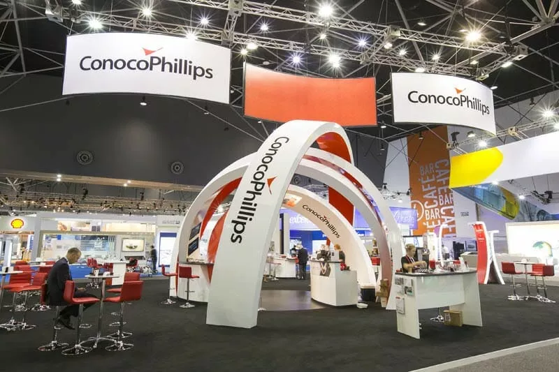 ConocoPhillips trade show booth with white arches and red accents, featuring company logos. Attendees in business attire engage at tables and bars.