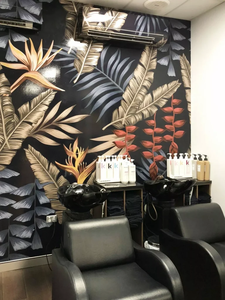 Two black salon chairs face washing sinks, set against a vibrant tropical leaf-patterned wall. Shelves hold white bottles. Sleek, modern vibe.