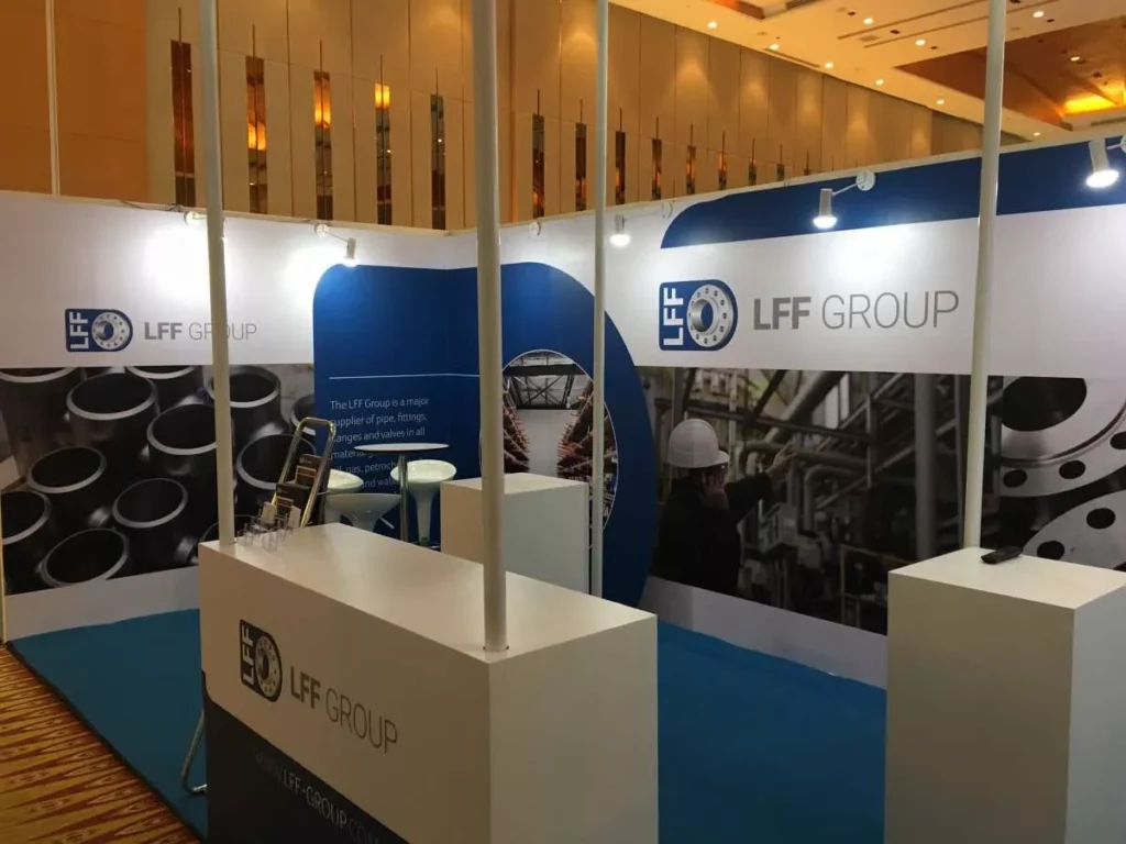 Exhibition booth with blue and white design, displays "LFF Group" logo. Background images of pipes and industrial worker in hard hat convey industry theme.