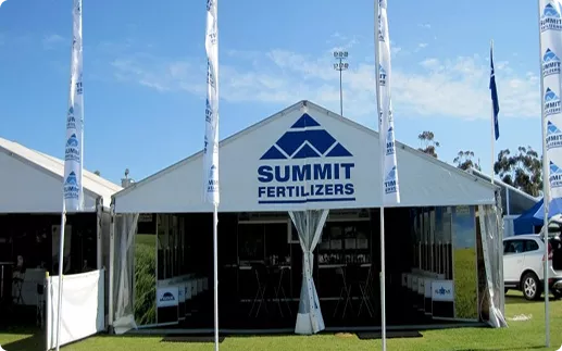 A white event tent with "Summit Fertilizers" branding stands on a grassy area under a clear blue sky. Tall banners flank the tent's entrance.