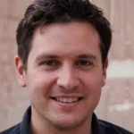 A smiling man with dark hair and a black shirt stands against a blurred neutral background. He appears relaxed and approachable.