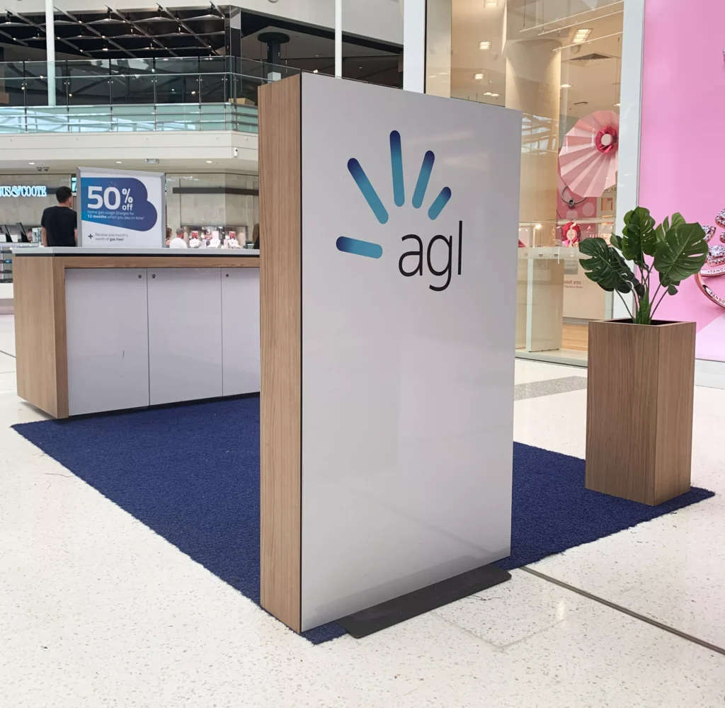 AGL promotional booth in a shopping mall with a sleek, white display panel featuring the AGL logo. A potted plant adds a touch of greenery