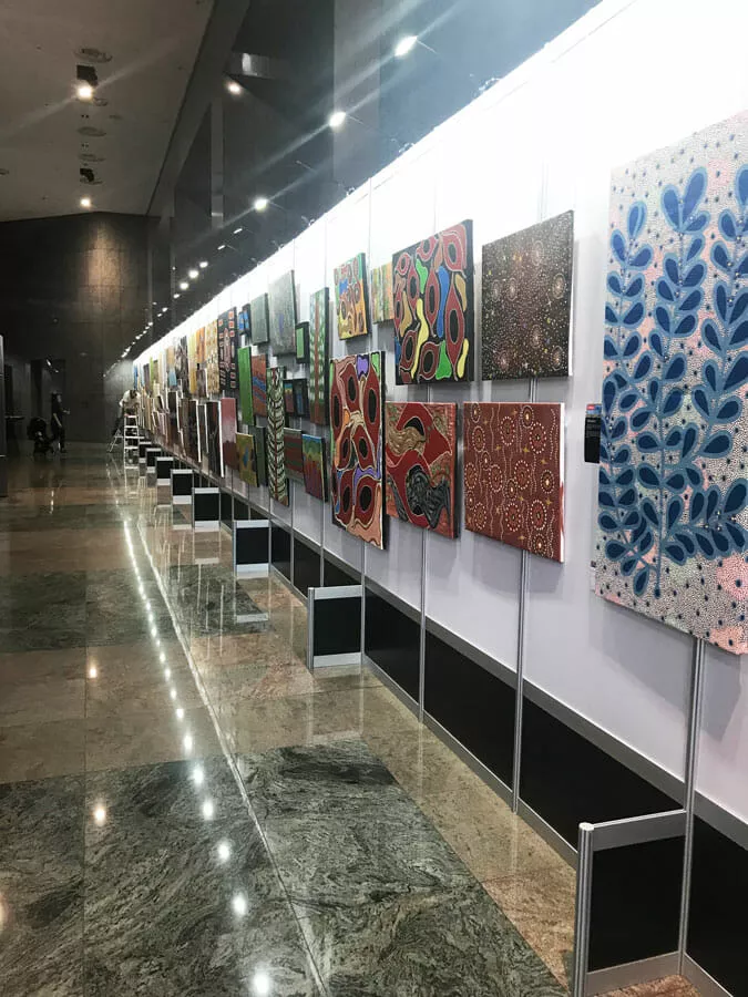 A gallery hallway with a long row of colorful abstract paintings on a white wall. The floor is shiny marble, and the space feels modern and bright
