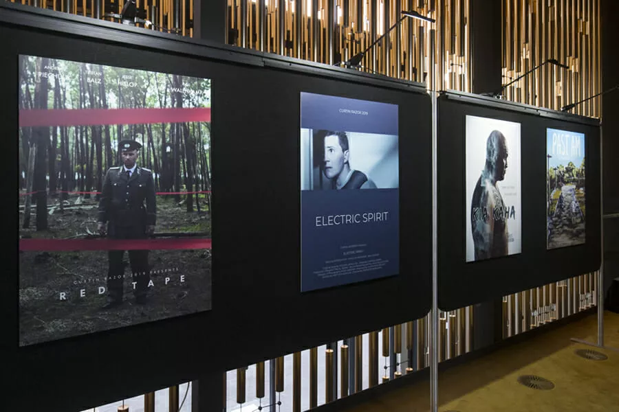 A series of four movie posters are displayed on a black panel in a well-lit room. From left to right, the posters are titled "Red Tape," "Electric Spirit," "Ka Kaha," and "Pastman." The first poster shows a uniformed man in a forest with red tape, the second has a man looking sideways, the third features a silhouette of a person with tattoos, and the fourth displays a landscape scene. Vertical slats behind add a modern touch.