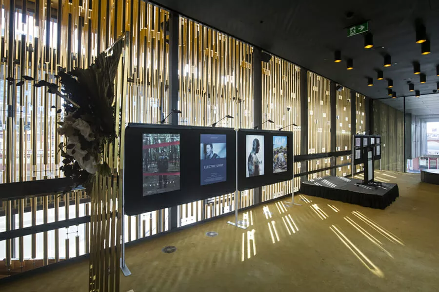 A modern gallery with vertical gold slats casting light patterns on the floor. Three display screens show artistic images; a chic, sophisticated atmosphere