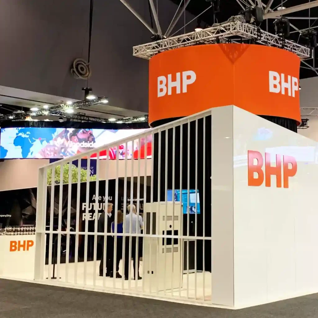 Exhibition booth for BHP with white and orange branding, featuring vertical slats and digital screens. Sleek, modern design at a trade show.