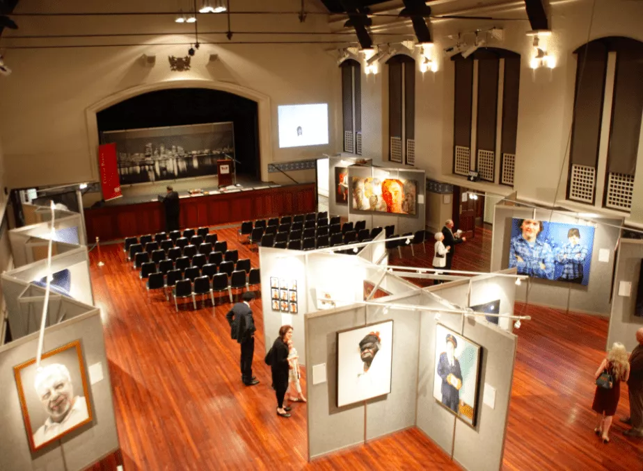 Art gallery interior with wooden floors, featuring portrait paintings on grey partition walls. A stage with a screen and empty chairs awaits an audience.