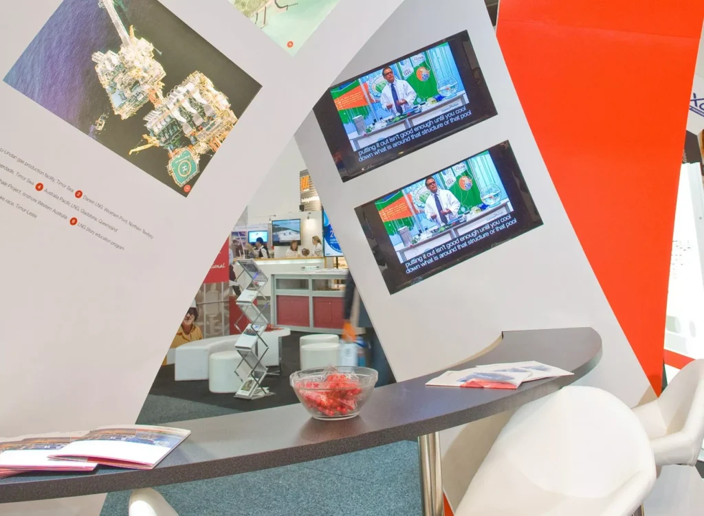 Curved exhibition wall with two monitors showing news anchors. Nearby, a curved table with brochures and a bowl of red candies
