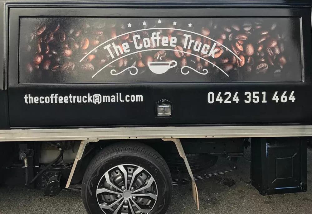 Close-up of a black coffee truck with a logo featuring coffee beans and a steaming cup. Includes contact details: email and phone number