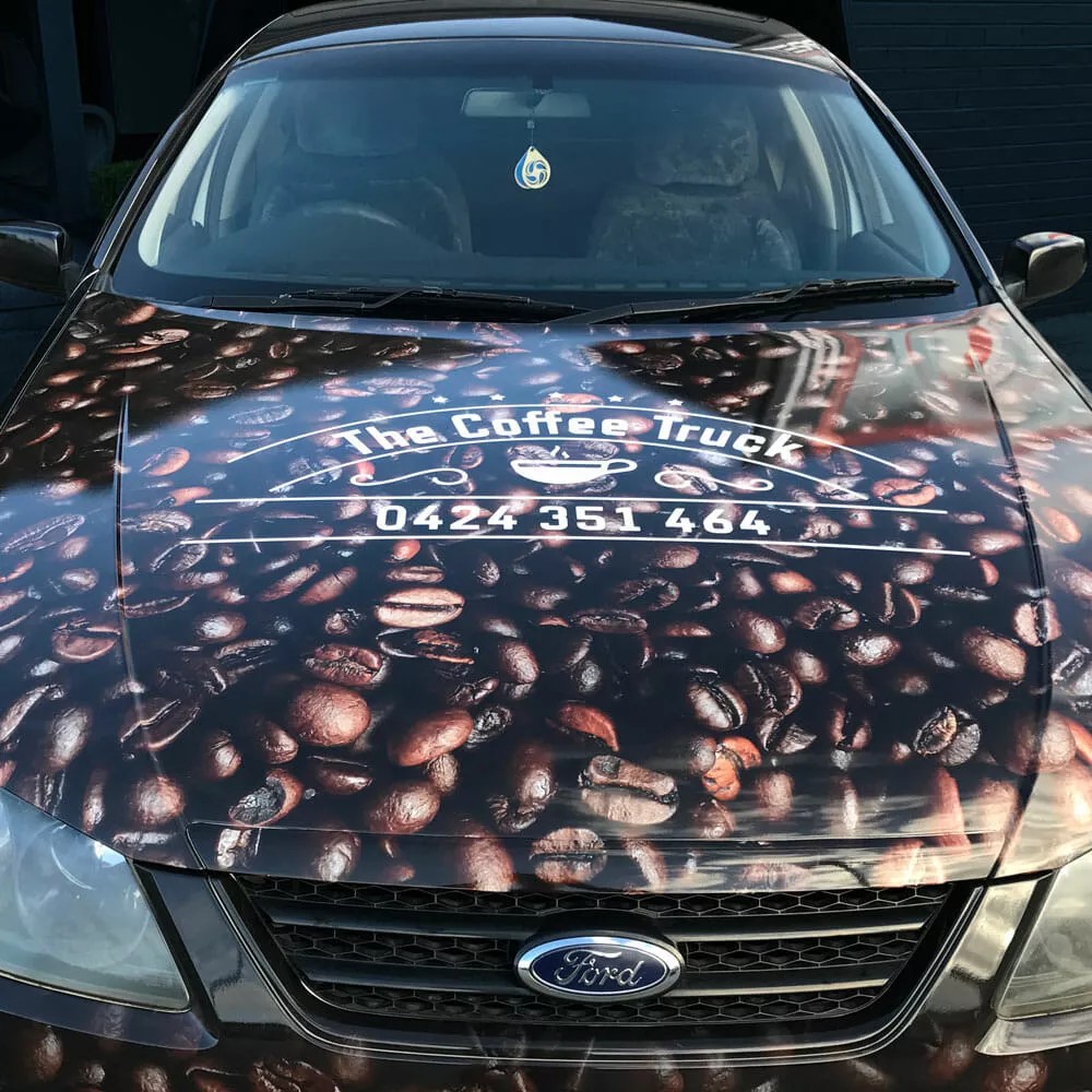 Close-up of a car hood with a coffee bean design, featuring the text "The Coffee Truck" and a phone number, providing a vibrant, caffeinated theme
