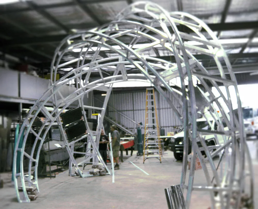 A large, intricate metal framework structure under construction in an industrial workshop. Workers and a ladder are visible, suggesting active progress.