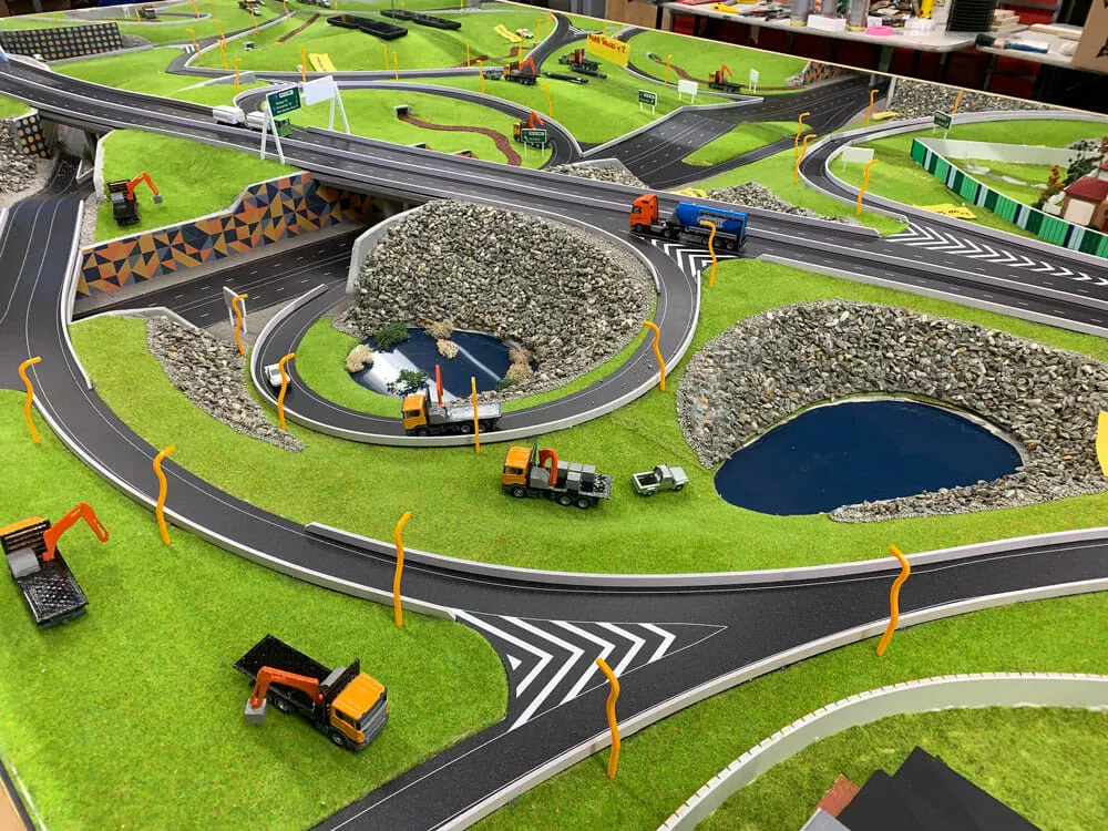 A detailed model landscape with winding roads, hills, bridges, and small construction vehicles on grassy terrain. Ponds surrounded by rocks add realism.