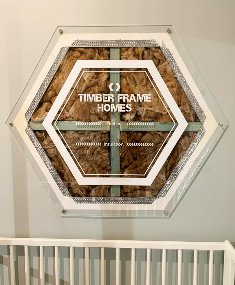 Hexagonal display labeled "Timber Frame Homes" shows wall insulation layers. Set on a light wall above a white railing.