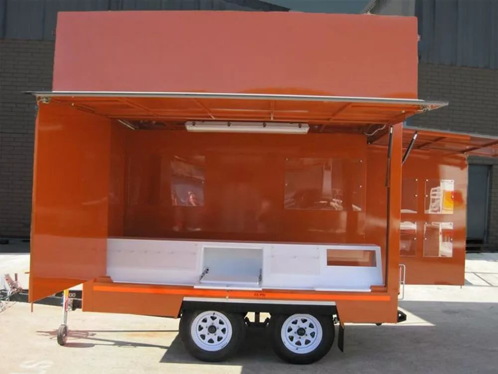 Open orange food trailer with dual axle wheels parked in front of a brick wall. The serving area and interior are empty, awaiting setup