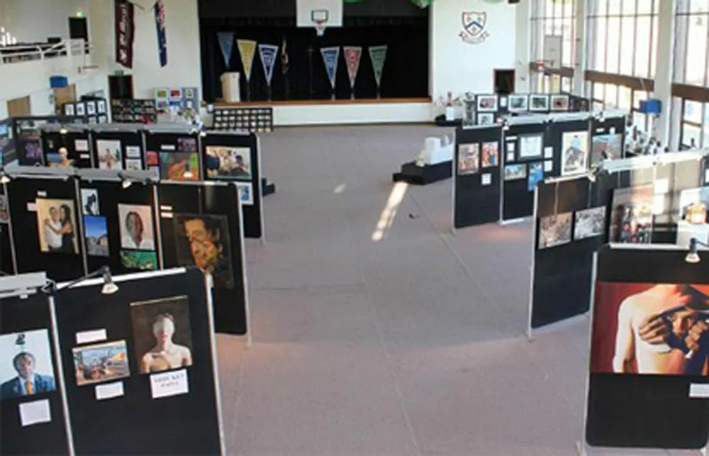 Art exhibition in a large hall with black display panels showcasing diverse artworks like paintings and photos. Sunlight streams in through tall windows, creating a warm, inviting atmosphere