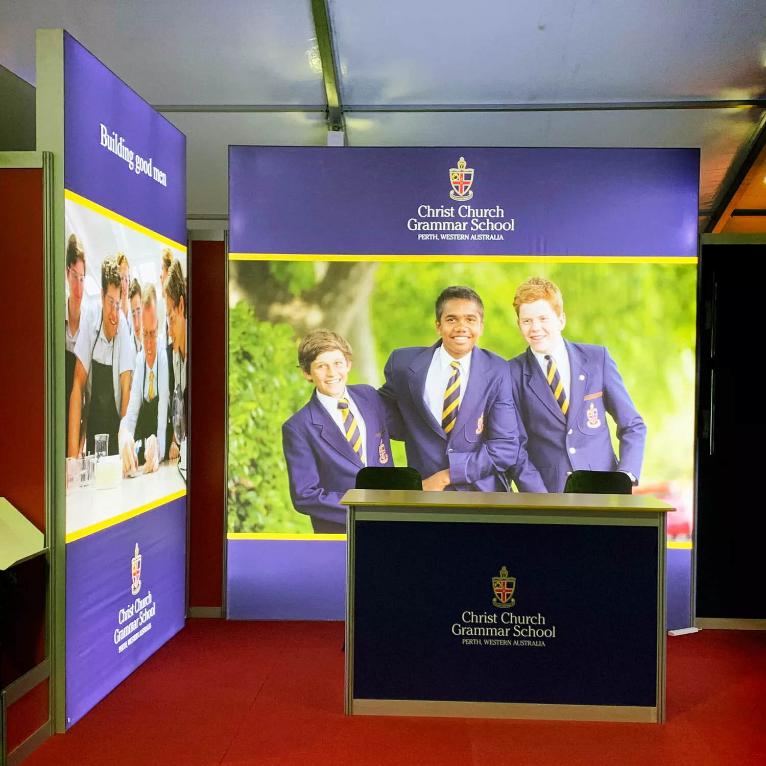 Booth for Christ Church Grammar School with a desk and backdrop featuring three students in blue blazers and ties, smiling in a green outdoor setting.