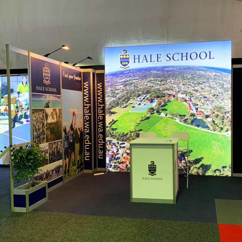 Exhibition booth for Hale School with a large aerial photo backdrop, a front counter with the school crest, side banners, and a potted plant.