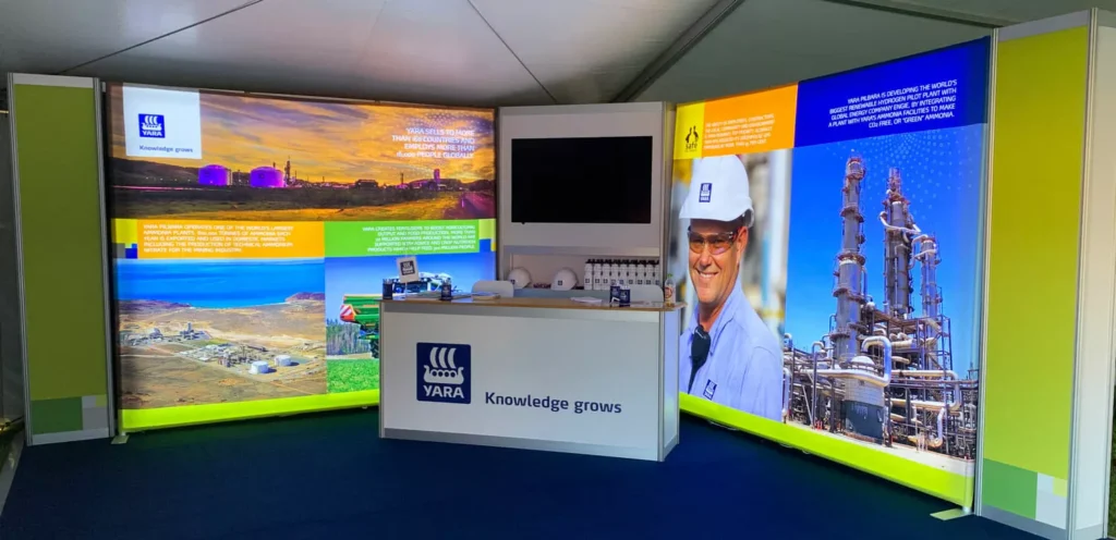 Exhibition booth with vibrant graphics depicting landscapes and industrial scenes. A smiling worker appears on the right. Central counter displays the logo.