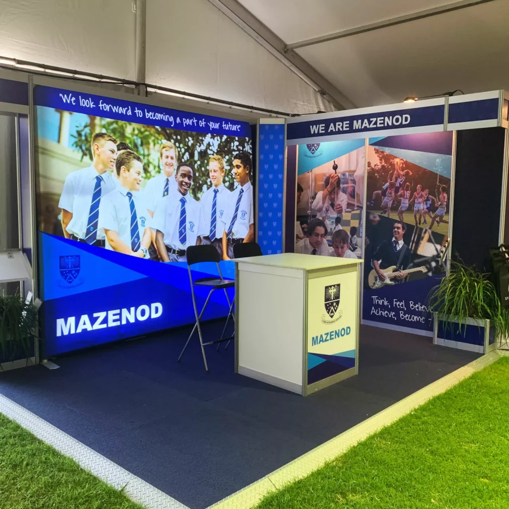 Exhibition booth featuring the Mazenod school logo, a smiling group of students in uniforms, promotional text, and colorful images.