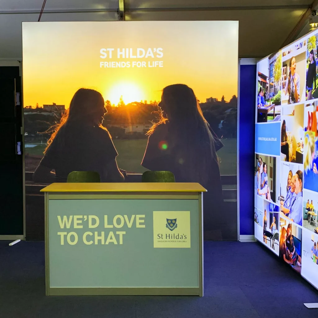 Exhibition booth for St Hilda's with sunset backdrop showing two silhouetted people, "Friends for Life" signage, and a counter reading "We'd Love to Chat".