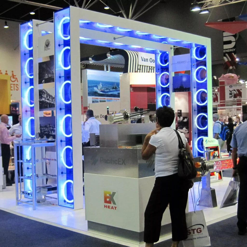 A trade show booth with a glowing blue LED frame features a person viewing displayed items. The setting is busy.