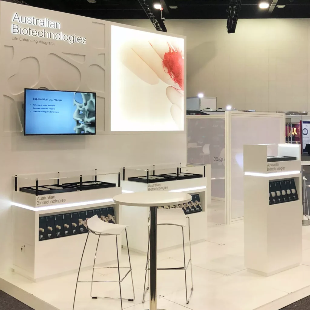 A sleek, modern biotech exhibition booth with a bright white design. Displays include a screen, decorative panels, and information counters.