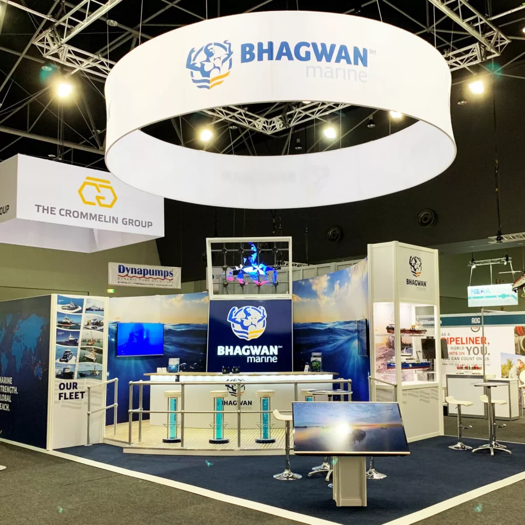 A well-lit exhibition booth for Bhagwan Marine, featuring branding, large circular overhead banner, digital screens, and seating arrangements. Sleek and professional tone.