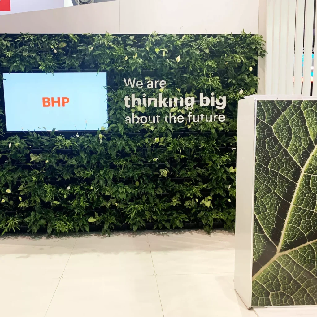 A green wall with lush foliage displays a screen showing "BHP" and the text "We are thinking big about the future." Nearby is a panel with a leaf pattern.