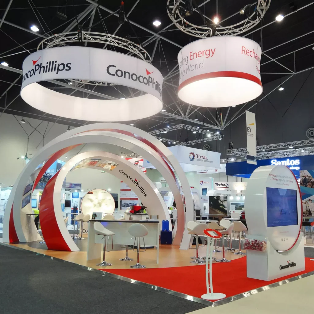 Exhibition booth with modern design showcases ConocoPhillips branding. Features a circular arch, interactive screens, and a red carpet, conveying innovation.