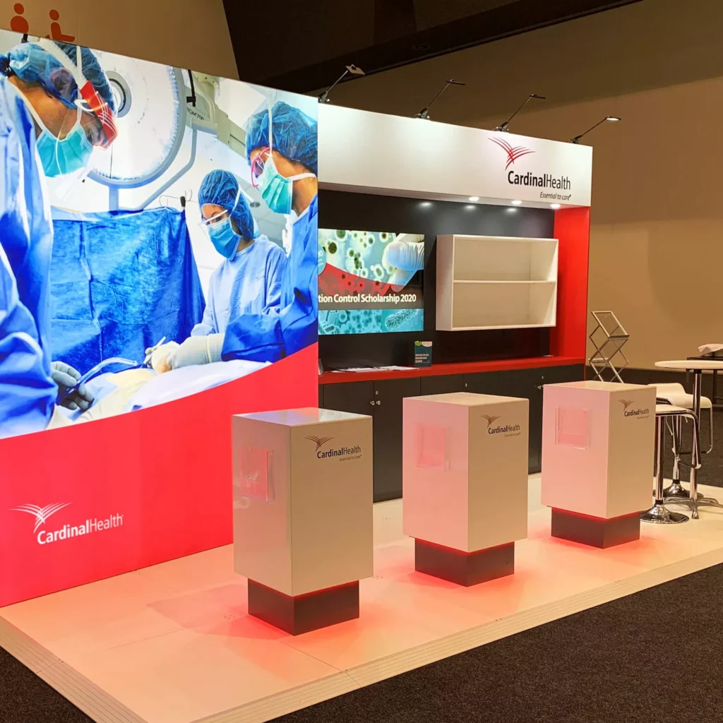 A trade show booth displays a large screen with surgeons in blue scrubs. Three white podiums are in front of a backdrop with the Cardinal Health logo.