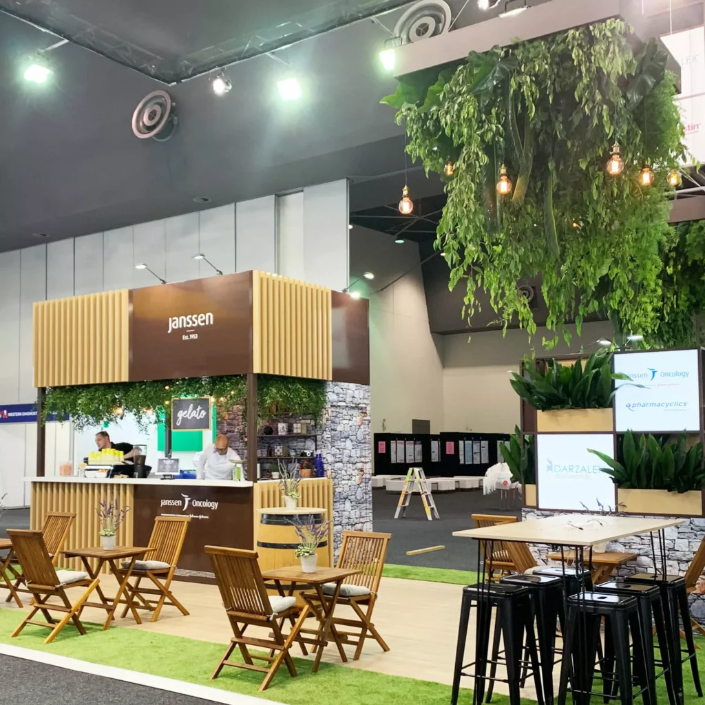 Modern exhibition booth with wooden accents, hanging plants, and Edison bulbs. Folding chairs and tables create a welcoming atmosphere.
