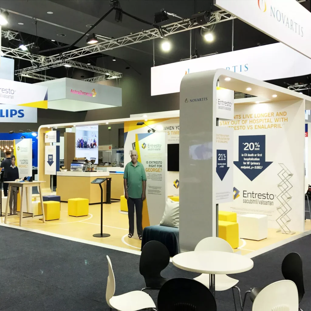 Modern exhibition booth by Barrett Exhibition Group with various seating areas and display stands, featuring yellow accents and overhead signage.
