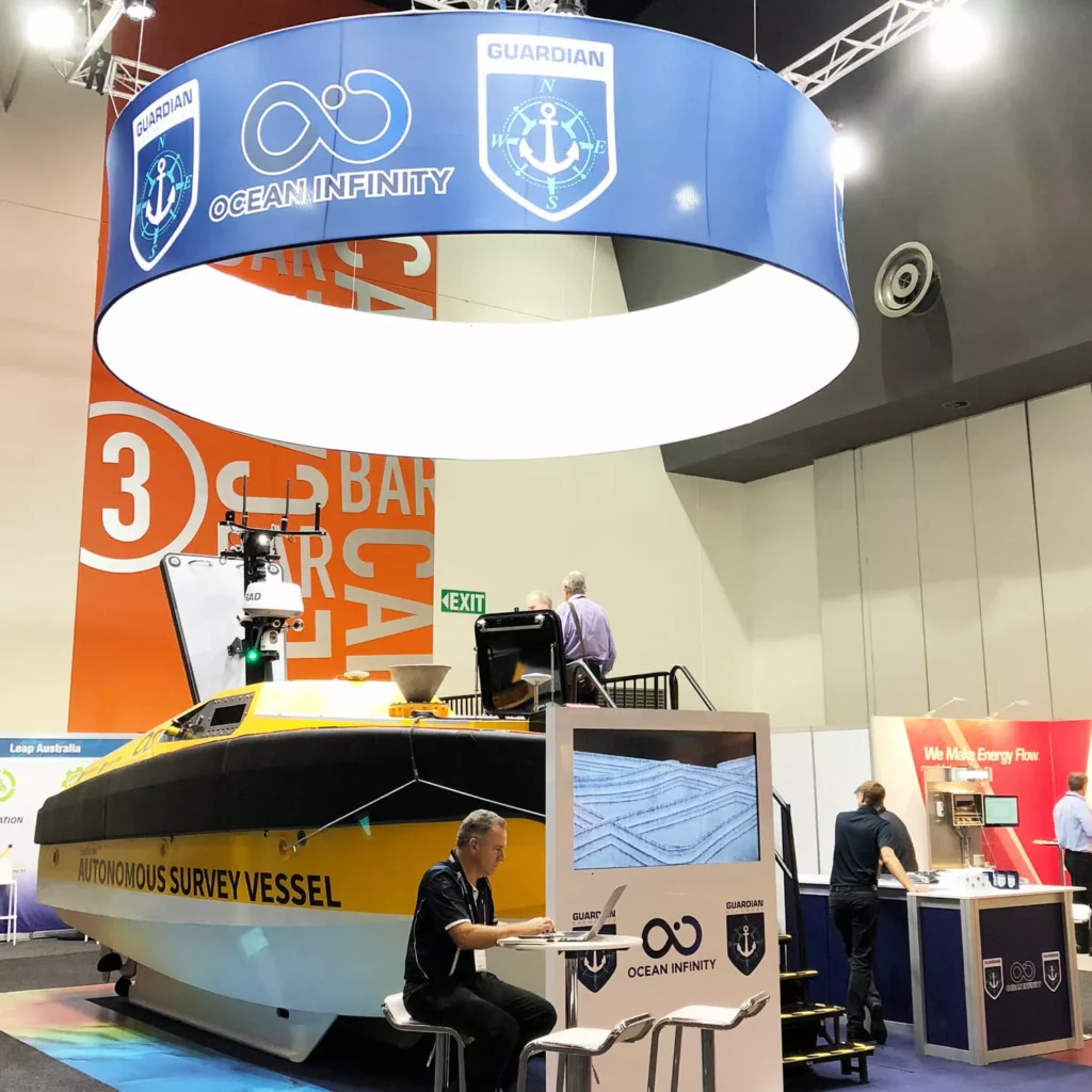 Exhibition booth by Barrett Exhibition Group featuring a model of an autonomous survey vessel, a large circular "Ocean Infinity" banner, and people engaging with displays.