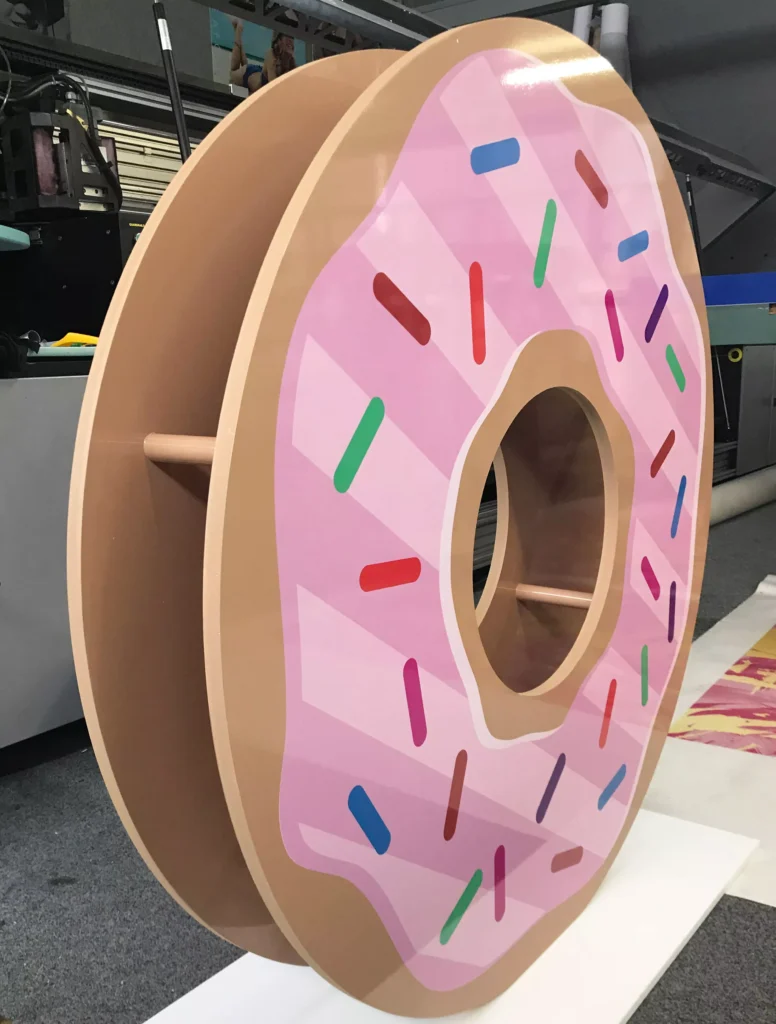 Large donut sculpture with pink icing and colorful sprinkles, featuring a hollow center. Set in a workshop, the mood is playful and whimsical
