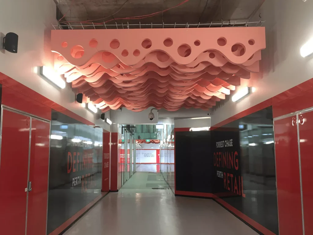 Corridor with a pink, wavy perforated ceiling installation. Walls are red and black with glass panels and retail advertisements. Modern, artistic vibe