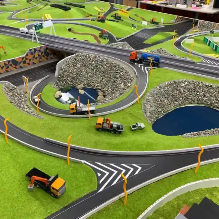 Model landscape with winding roads, bridges, small trucks, and vibrant green grass. Features rock formations and blue water pools, creating a lively scene.
