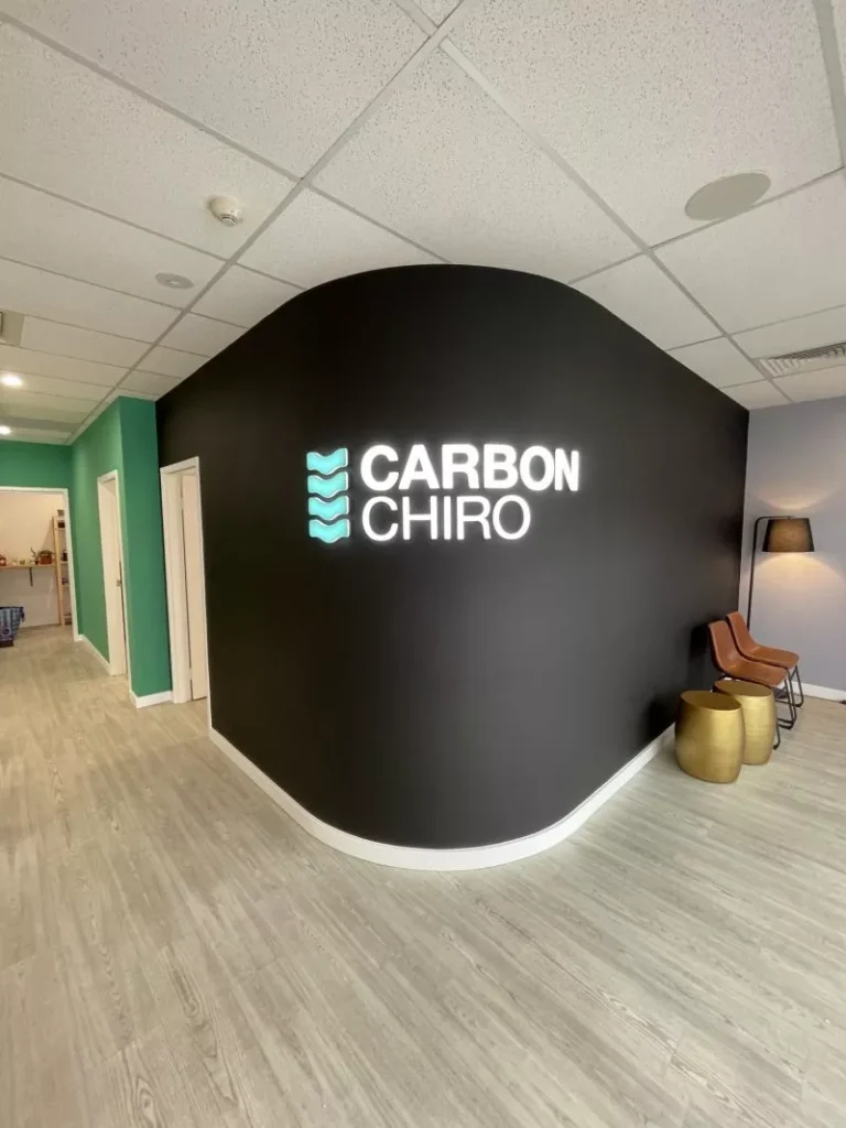 Modern chiropractic office with light wood flooring, teal walls, and a curved black feature wall displaying "CARBON CHIRO" in glowing white letters