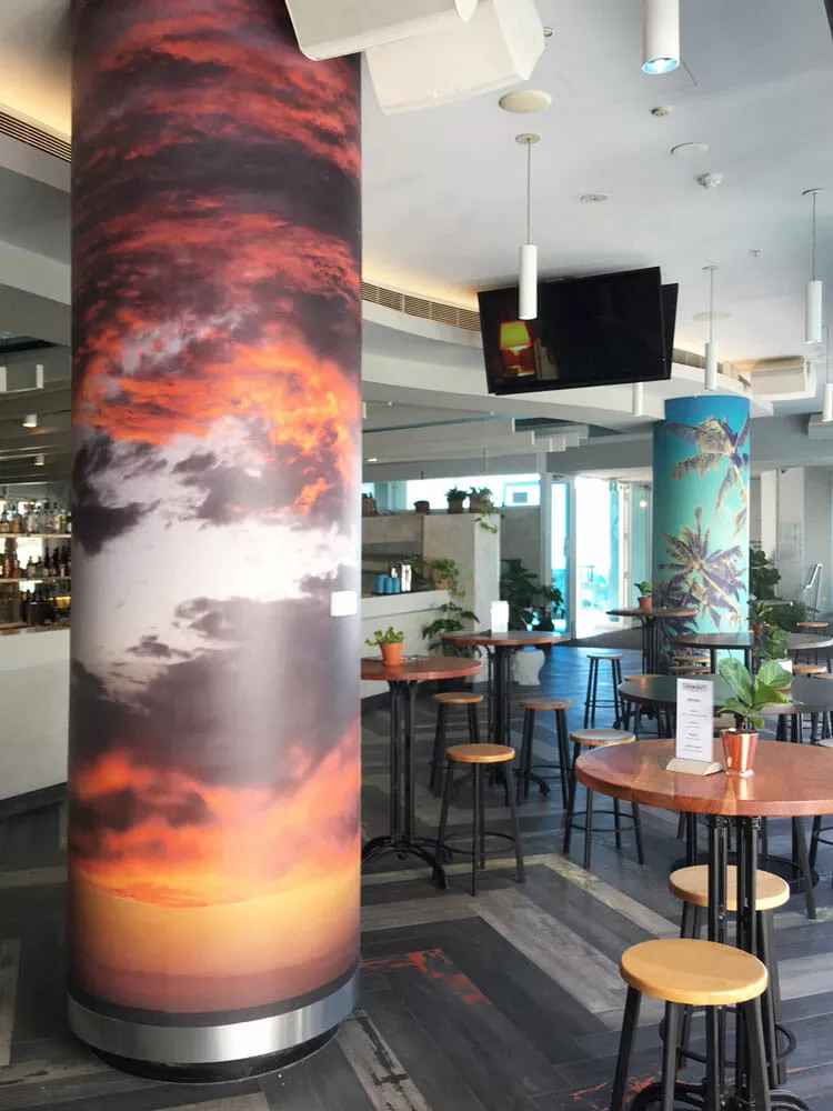 A modern café interior featuring tall columns wrapped in vibrant sunset and palm tree images. High tables with wooden stools and scattered potted plants create a relaxed atmosphere