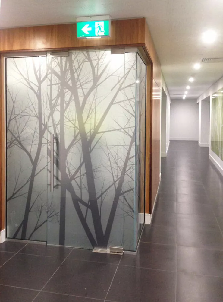 Glass office doors with a tree branch design in frosted pattern, under an exit sign. A long, well-lit hallway with gray tile floors extends to the right