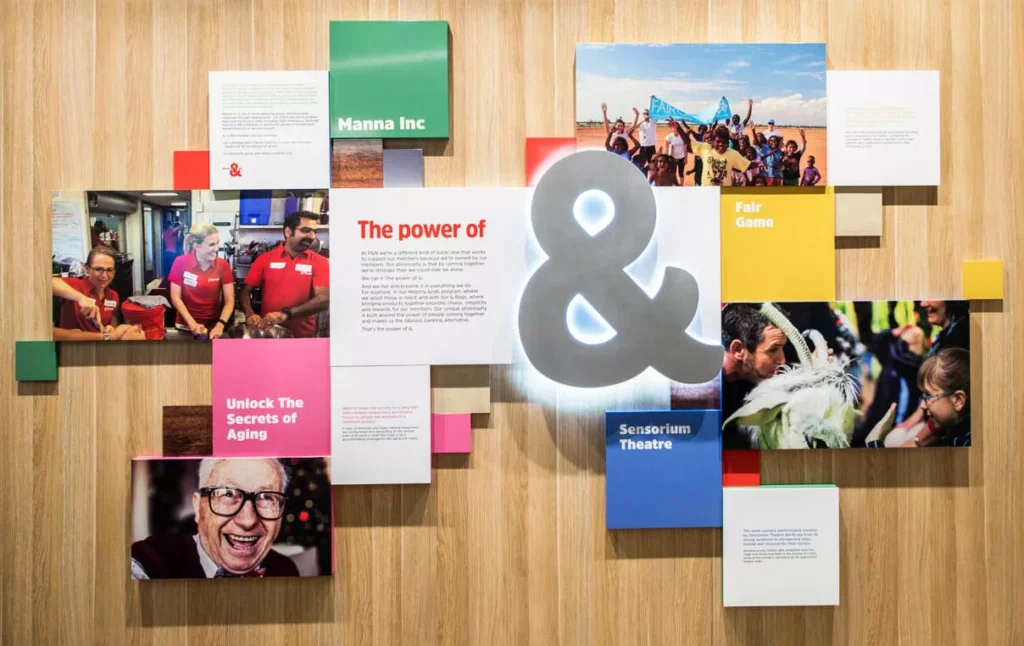 A collage of colorful panels on a wooden wall features vibrant photos of community activities, text, and a large illuminated ampersand. Keywords include "Manna Inc," "Fair Game," and "Unlock The Secrets of Aging," conveying a sense of community and inclusivity